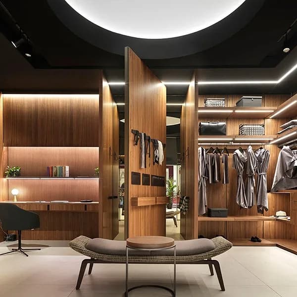 Barausse walnut pivot door opening onto a lit walk-in wardrobe, with a panelled study wall and a woven bench in the foreground