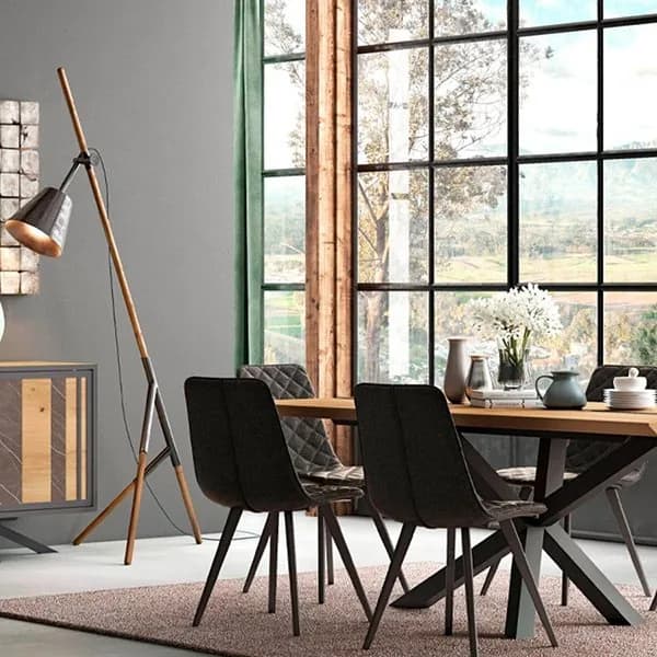 Clara & Home oak dining table on a black star base with dark quilted chairs, beside a wooden tripod floor lamp and a full-height black-framed window overlooking countryside